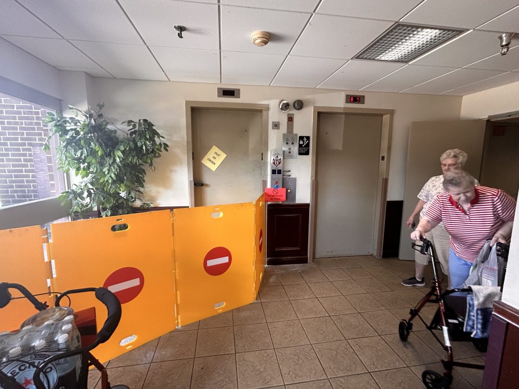 Two women, one leaning on a walker, stand in a hall near two elevators. One elevator with an out of order sign on the door has four barriers in front of it.