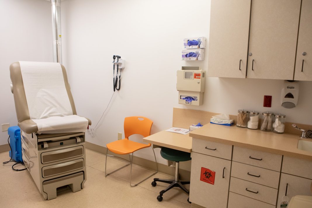 A clinical examination room with a brown examination table, orange chair, medical equipment on the wall, and a cabinet holding medical supplies.